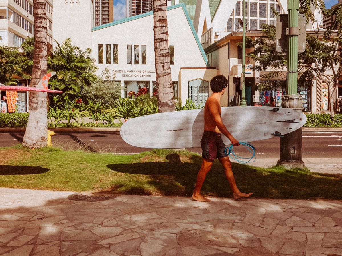 A person walking barefoot on a sidewalk carrying a surfboard; palm trees and buildings are in the background.