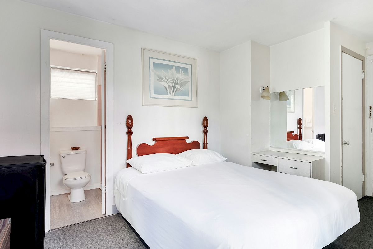 Bright bedroom with a large bed and vanity mirror, plus an open door to a small bathroom with a toilet.