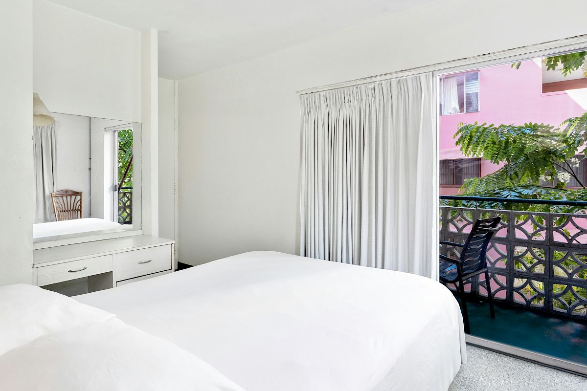 A bright bedroom with a white bed, built-in dresser, and sheer curtains opening to a balcony with chairs and greenery.