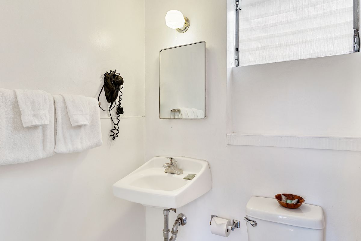 A clean hotel bathroom with a small sink, faucet, folded towels, mirror, light fixture, and a toilet with paper.