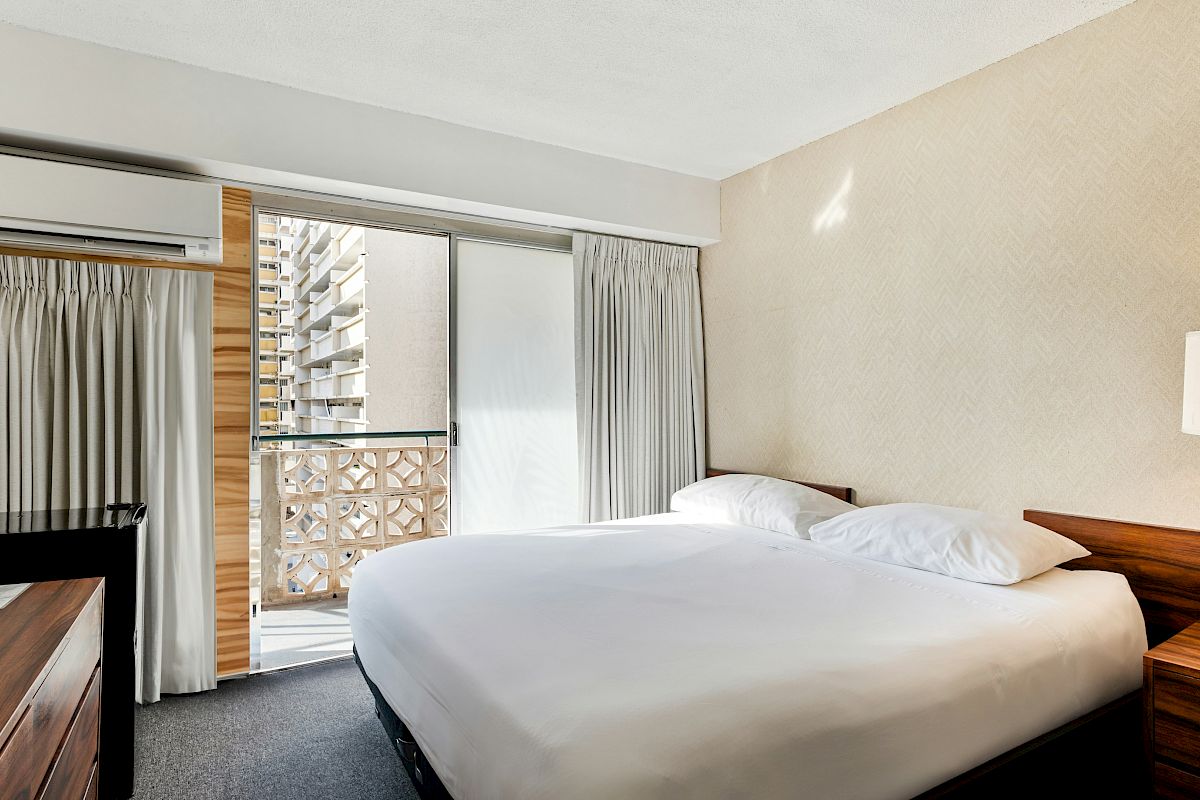 A hotel bedroom with a king bed, wall AC unit, gray curtains, and an open balcony door overlooking apartment buildings.