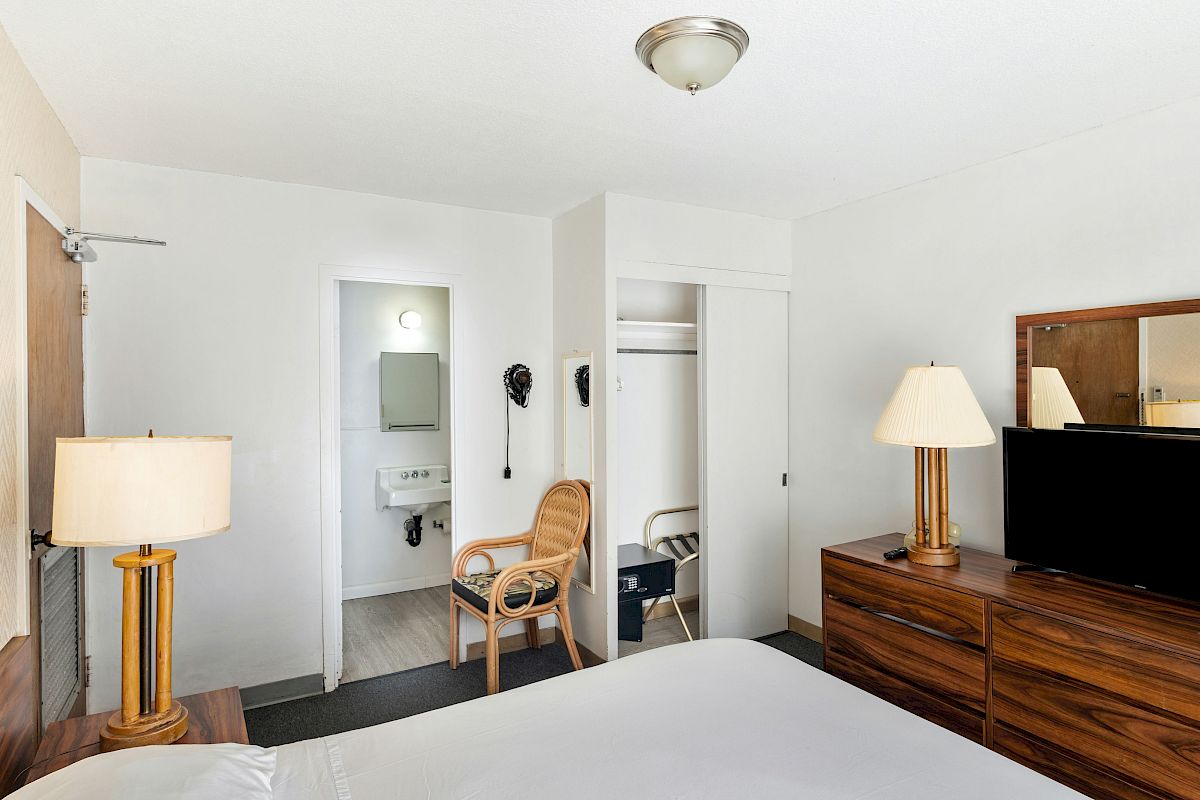 A bright hotel room with a TV on a wooden dresser, a lit lamp, a chair, and an open bathroom doorway with fixtures.