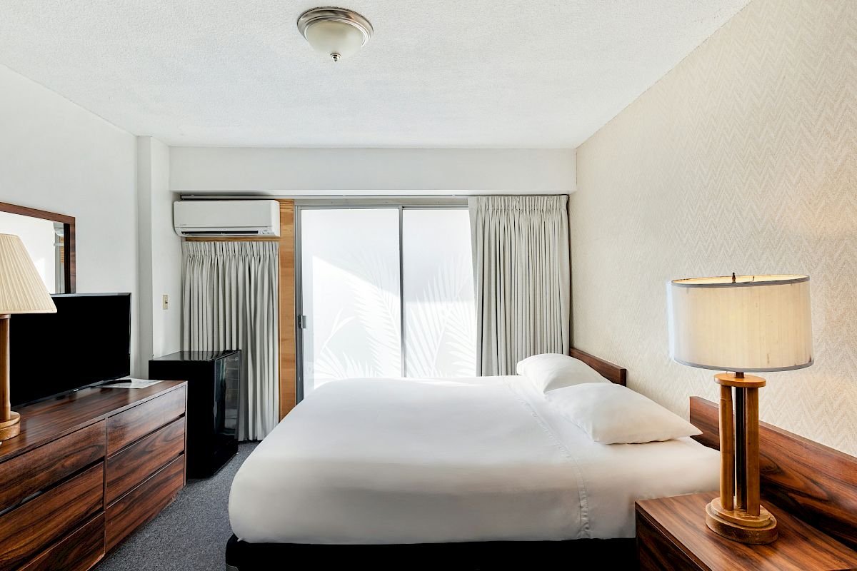 A hotel-style bedroom with a made bed, bedside lamps, a TV on a dresser, and an AC unit by a sliding door with curtains.