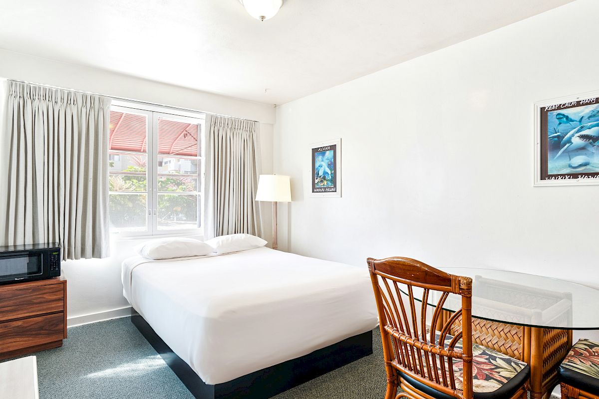 A bright small bedroom with a single bed, gray curtains, wall art of dolphins, a lamp, microwave, and a wicker table.