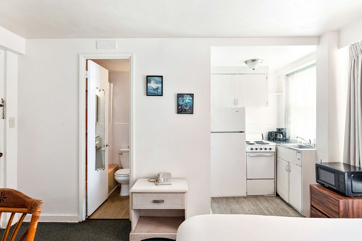 Bright apartment interior showing a compact kitchen with stove, sink and cabinets, plus a view into the bathroom and toilet.
