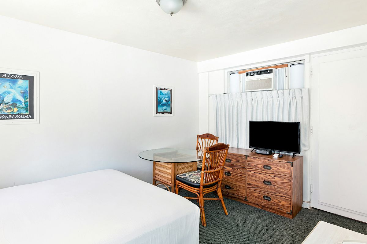A bright hotel room with a bed, glass table, rattan chair, TV and air conditioner, and “Aloha” wall art.