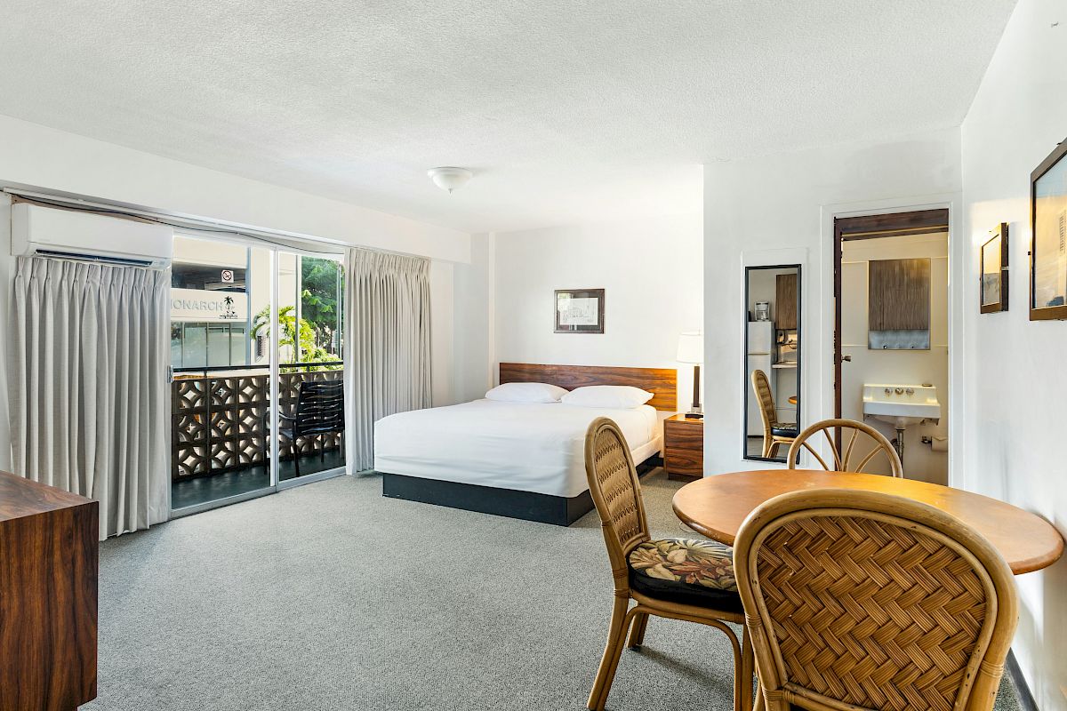 Bright hotel room with a bed, balcony doors, curtains, a small table with wicker chairs, and a vanity/sink area.