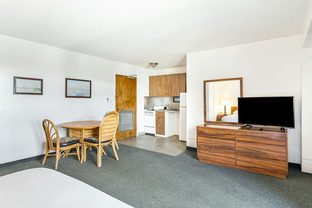 A bright hotel room with a dining table and chairs, a TV on a wooden stand, a kitchenette, and a view of the bed through an open doorway.