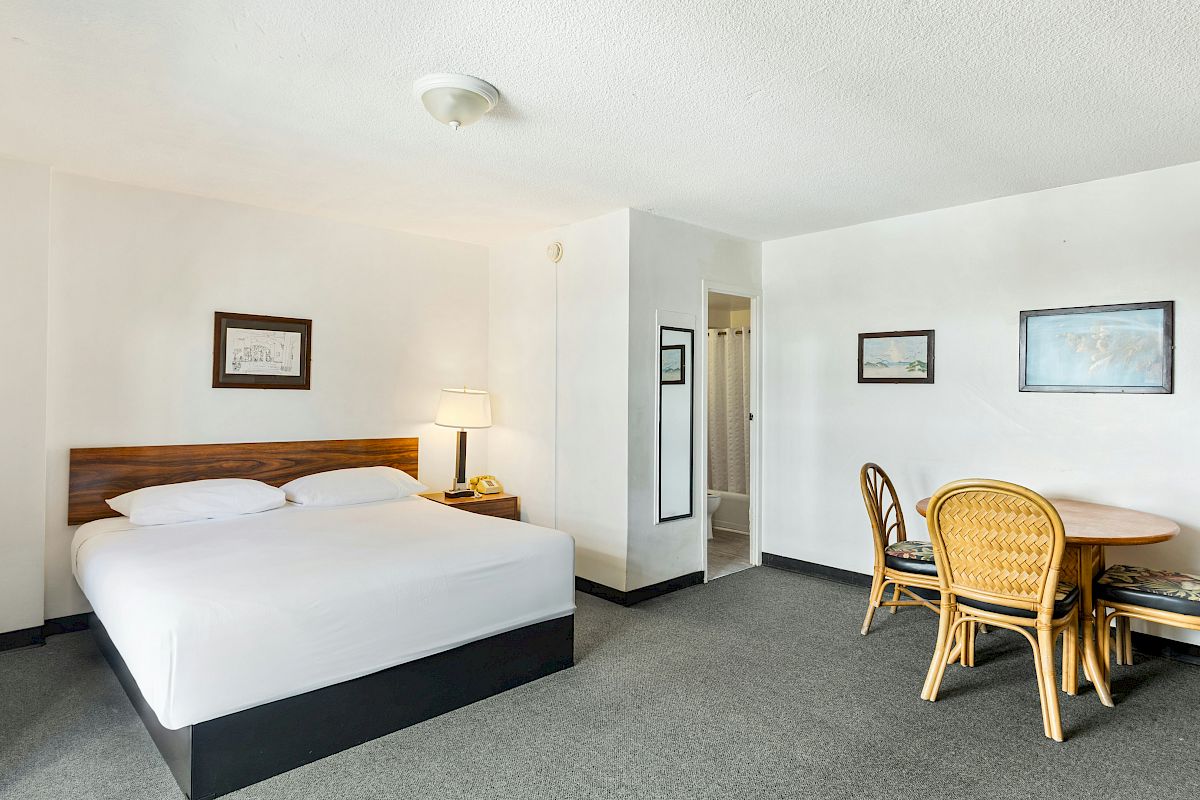 A bright hotel room with a bed, bedside lamp, small dining table, wicker chairs, framed pictures, and an overhead ceiling light.