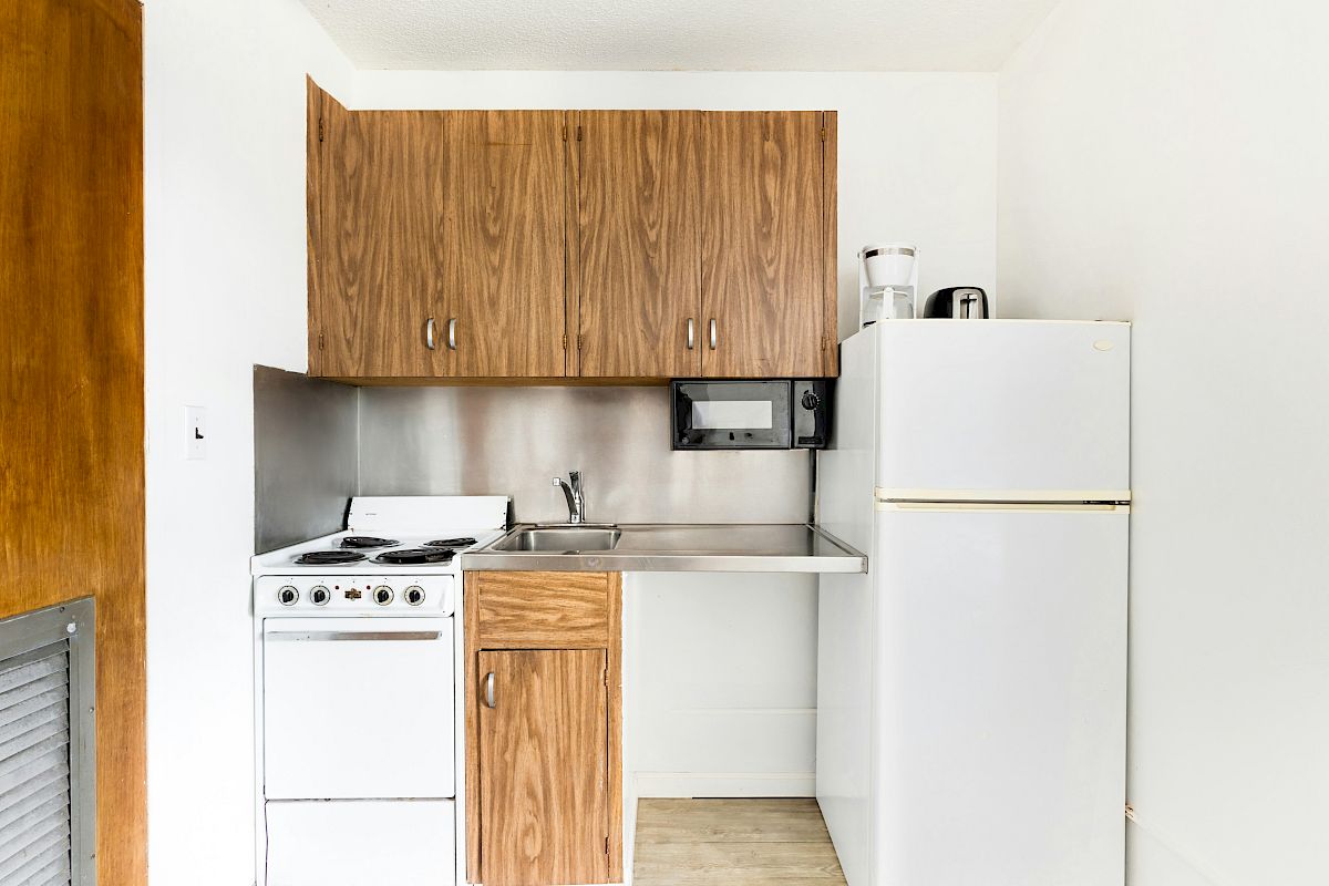 A compact kitchen with wood upper cabinets, a stainless backsplash, sink, stove, and a white refrigerator.