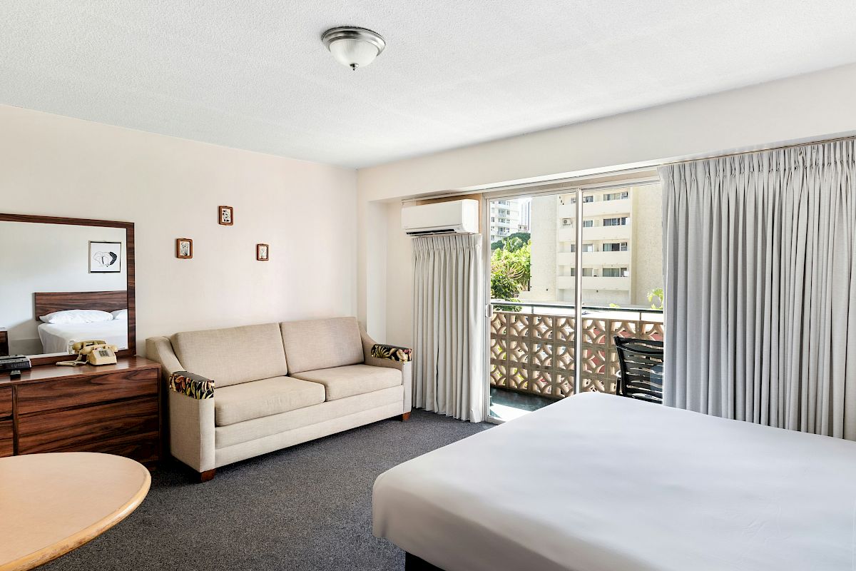 A tidy hotel room with a sofa, bed, window sliding doors, gray curtains, wall art, and an air conditioner on the wall, above.