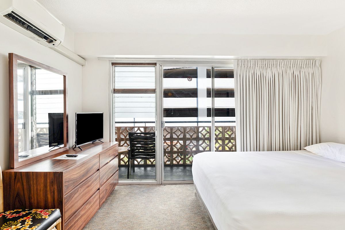Bright hotel bedroom with a bed, TV on a wooden console, air conditioner, beige curtains, and sliding doors to a balcony.