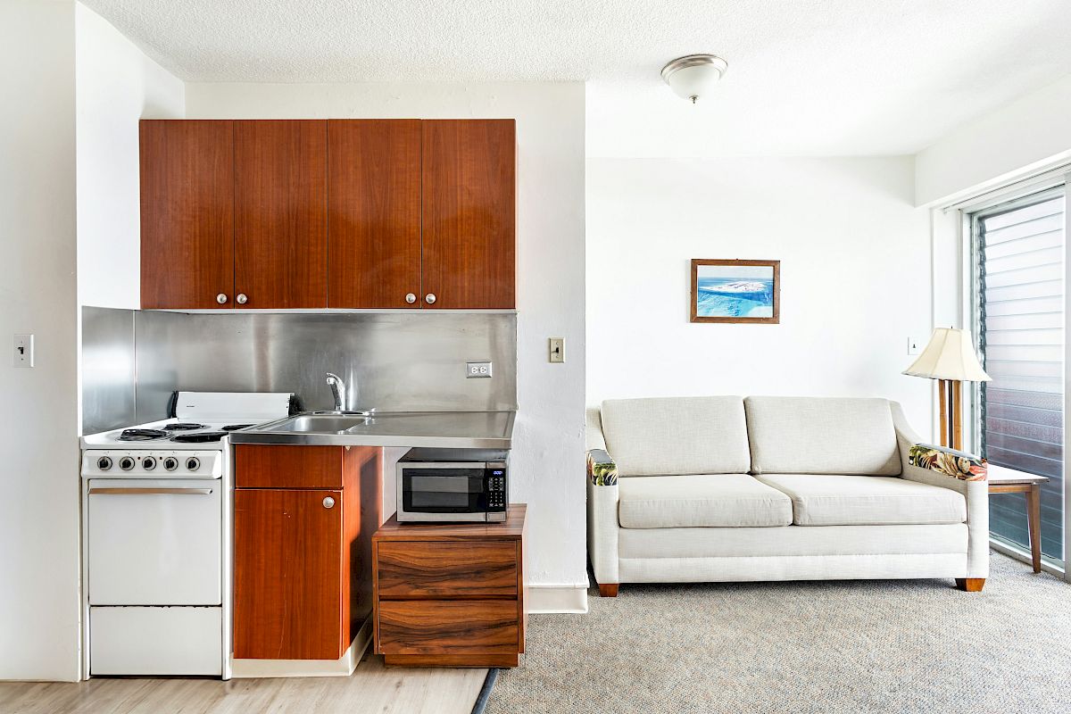 A small apartment kitchenette with wood cabinets, stove, sink, and microwave beside a cozy living room with a sofa.