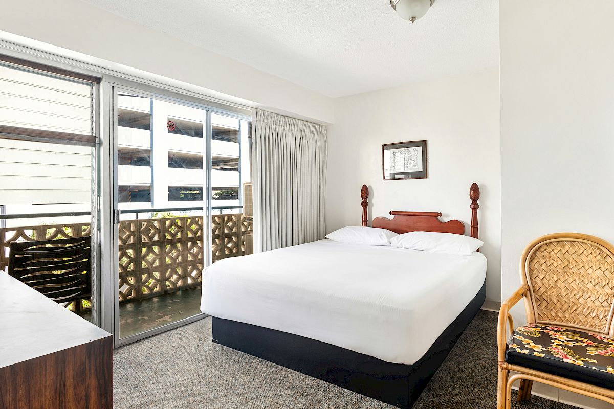 A bright bedroom with a wooden bed, open sliding doors to a balcony, a wicker chair, and framed wall art, all neatly staged.
