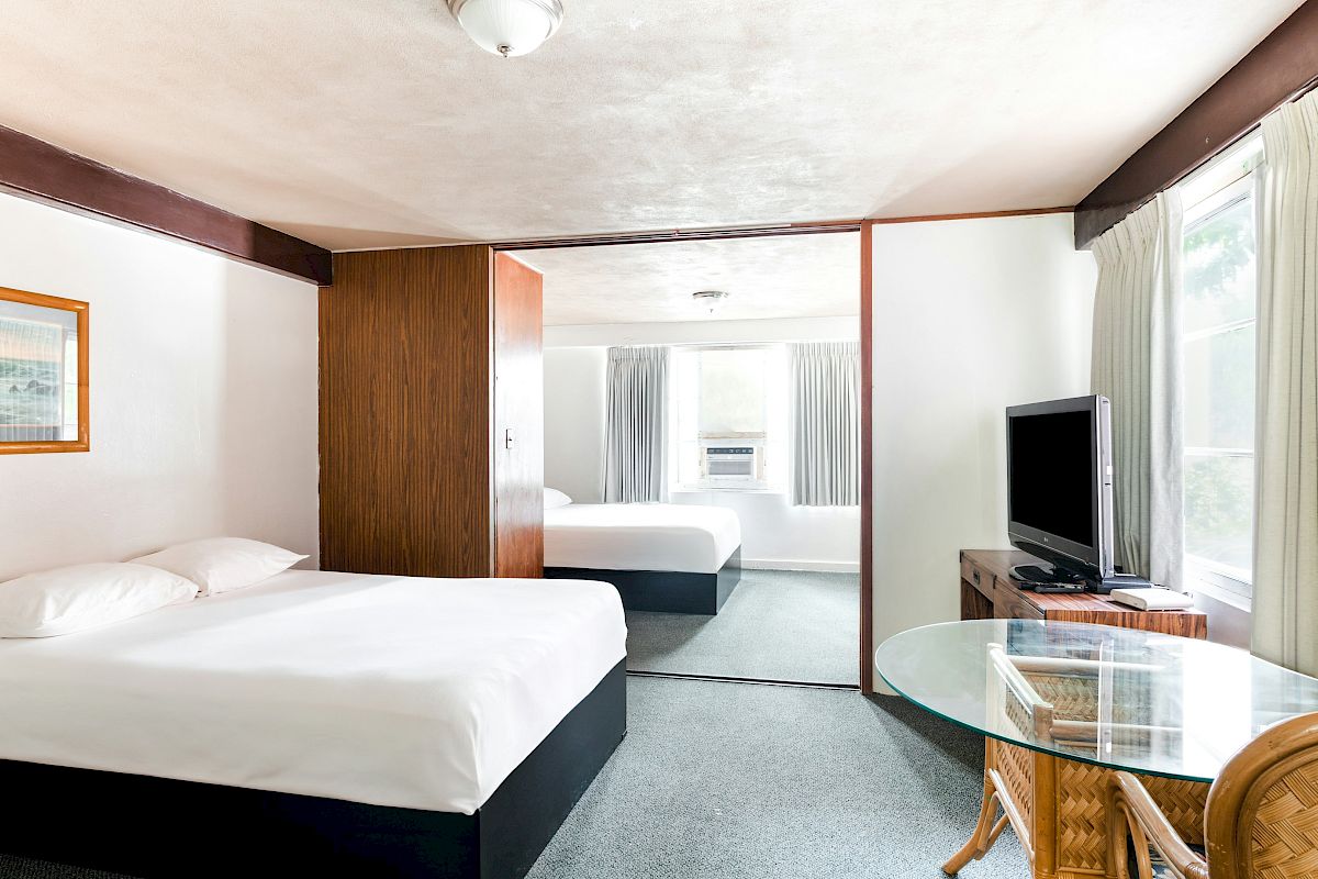 A bright hotel room with two beds, a wall-mounted TV, curtains, and a small glass-topped table in front of a window.