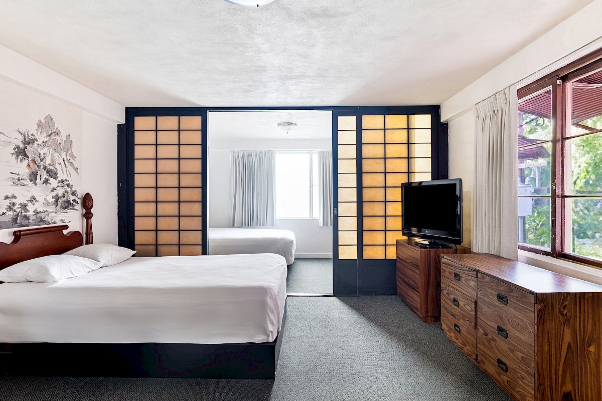 A modern hotel bedroom with a bed, TV on a wooden dresser, paneled wall, and large window with curtains.