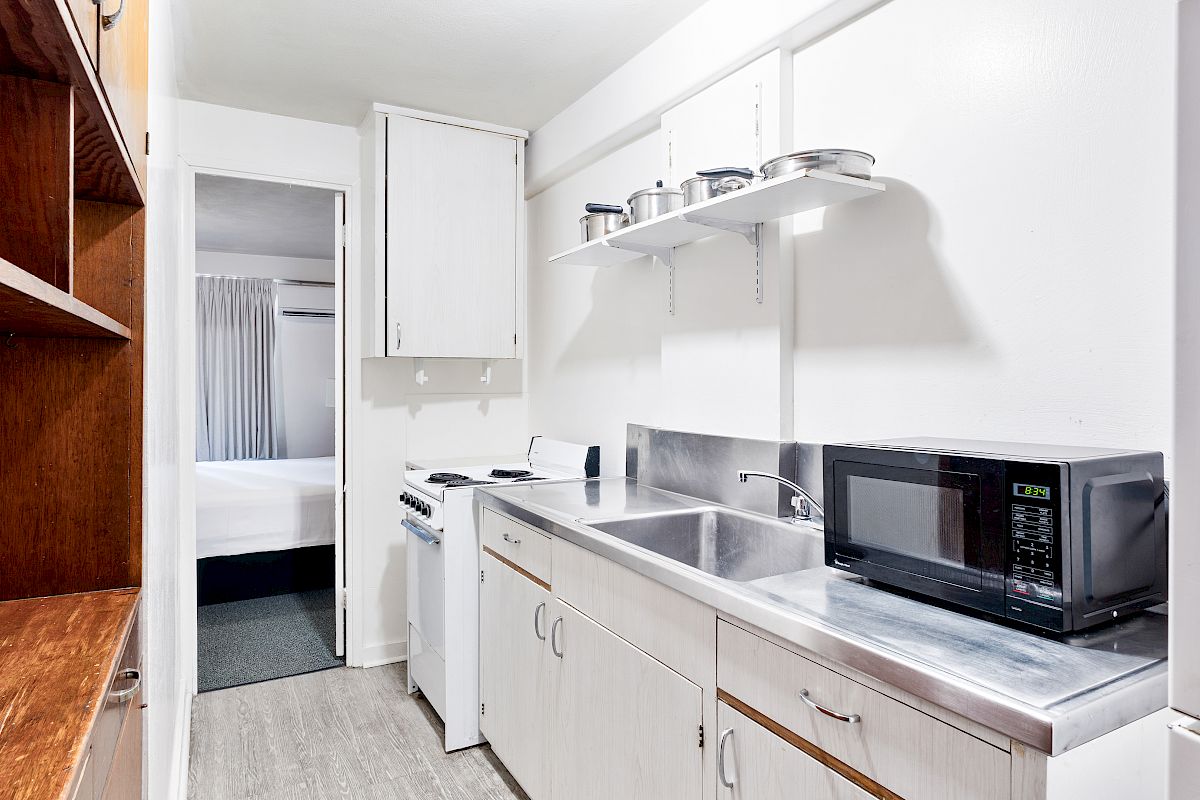 A bright kitchenette with a stove, sink, microwave, overhead pots, cabinets, and an open doorway to a bedroom.