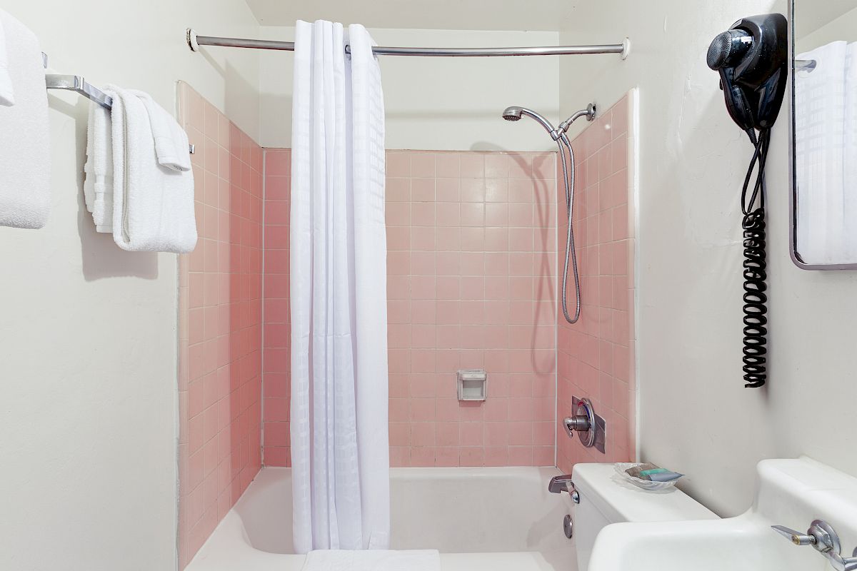 A small bathroom with a bathtub, white shower curtain, pink-tiled walls, folded towels, and a handheld shower head on a hose.
