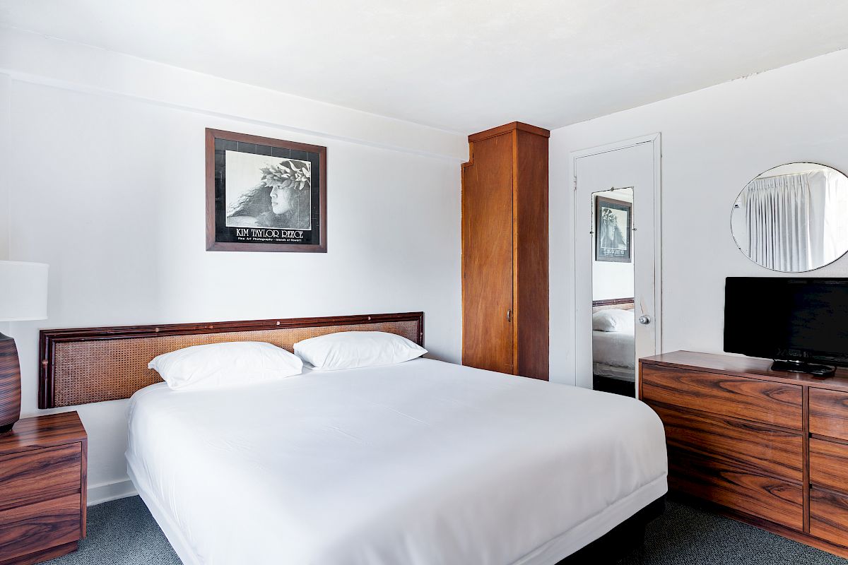 A tidy hotel bedroom with a large bed, wooden dresser, TV, mirror, framed art, lamp, and an open closet doorway.