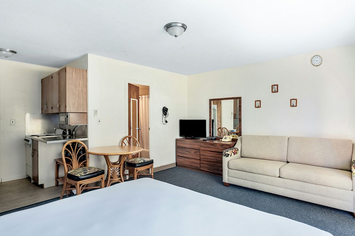 A bright living room with a beige sofa, TV on a wooden cabinet, a kitchenette, and a small dining table and chairs.