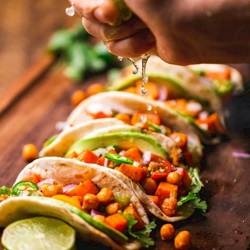 A hand squeezes lime over tacos filled with vegetables and chickpeas on a wooden board, with sliced lime in the foreground.