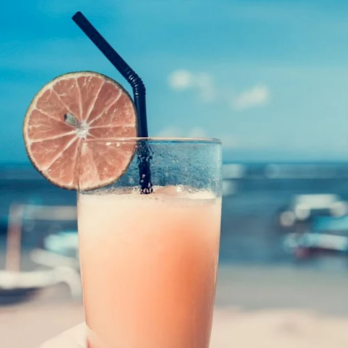 A refreshing drink in a glass with a black straw and a lime slice on the rim, held up with a beach and ocean in the background.