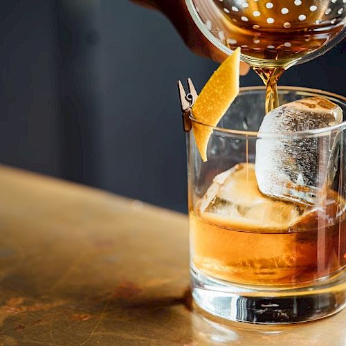 A drink being poured into a glass with ice and garnished with a twist of citrus peel, set against a dark background.