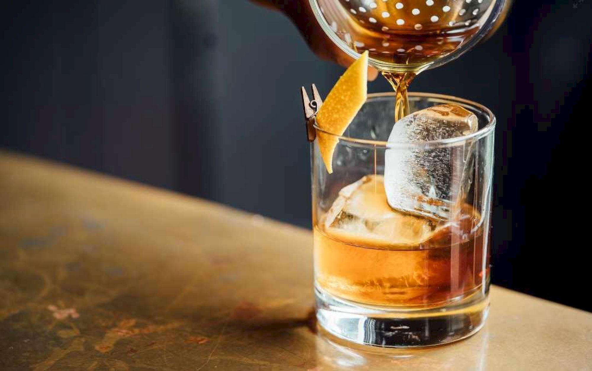 A cocktail being poured into a glass with ice and a citrus peel garnish on a wooden surface.