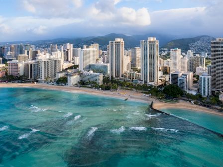A coastal cityscape featuring tall buildings, a sandy beach, and turquoise waters under a partly cloudy sky.