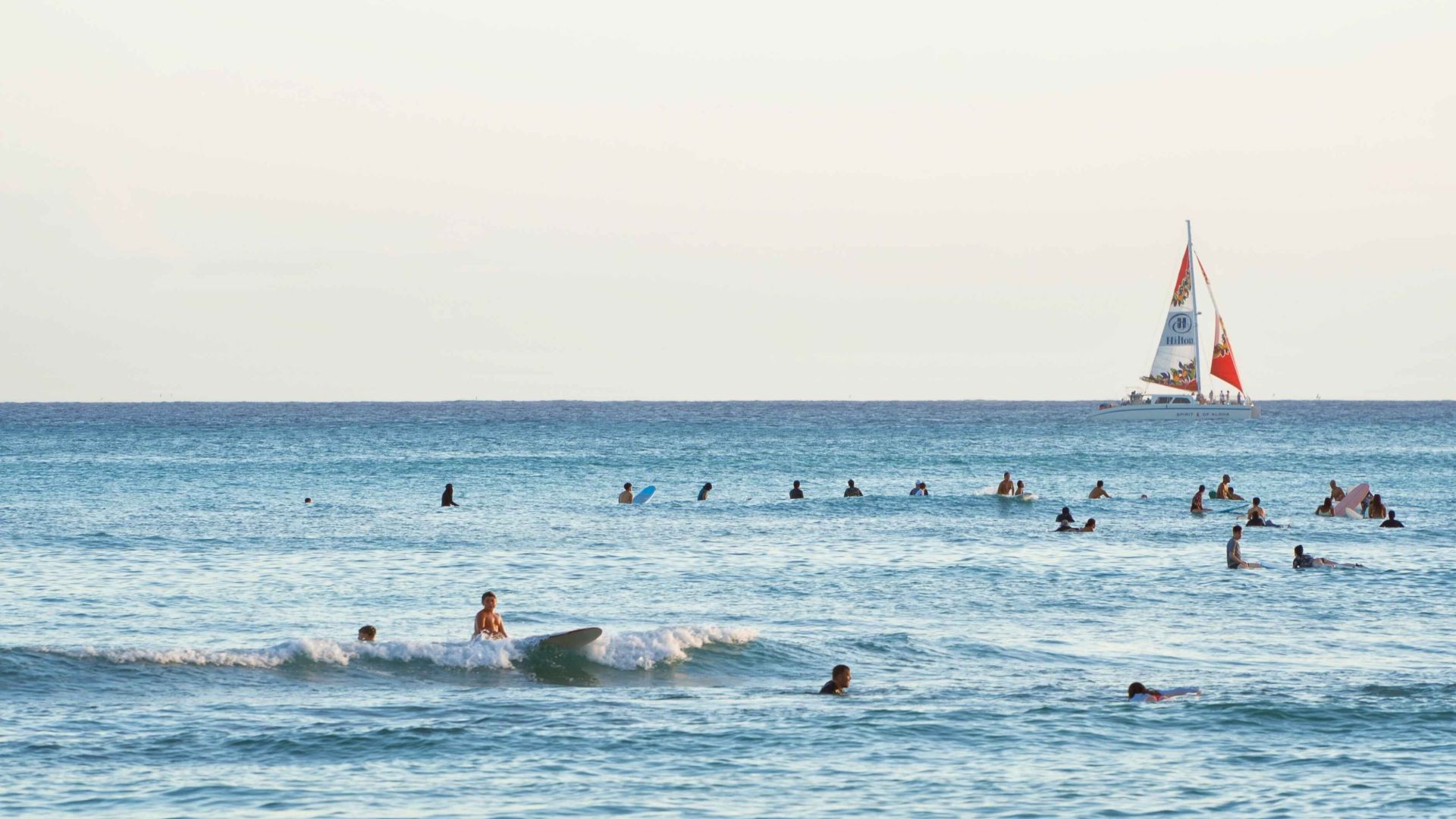 People are swimming and surfing in the ocean, with a sailboat in the distance on a clear day.