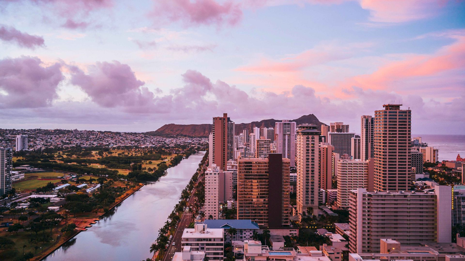 A cityscape at sunset features tall buildings, a river, and a mountain range under a pink and purple sky.