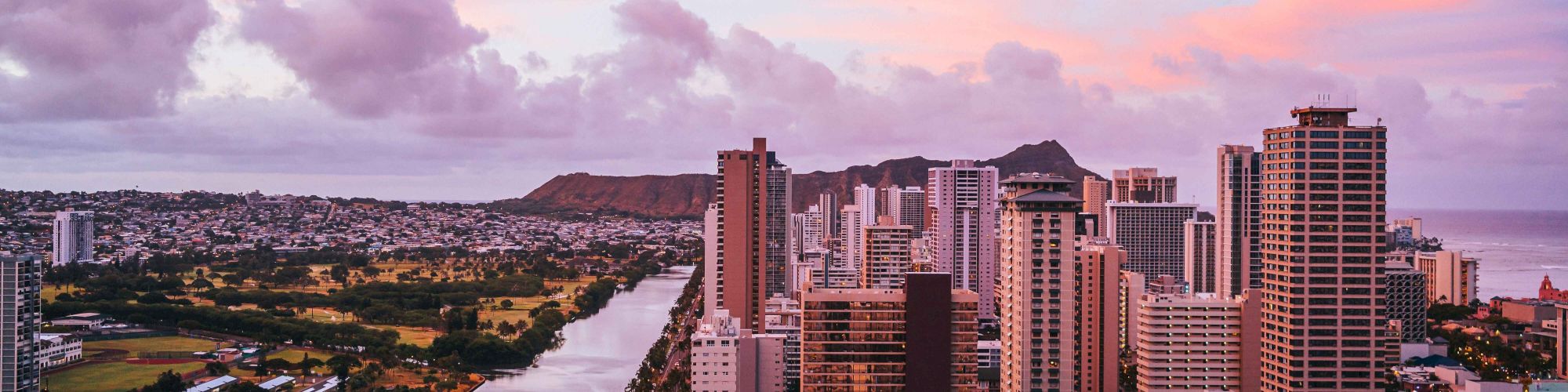 A cityscape at sunset features tall buildings, a river, and a mountain range under a pink and purple sky.