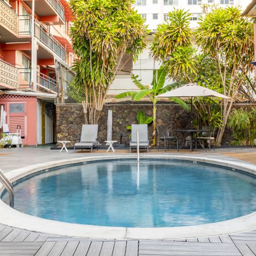 A circular pool surrounded by loungers, umbrellas, and tropical plants, with a multi-story building in the background.