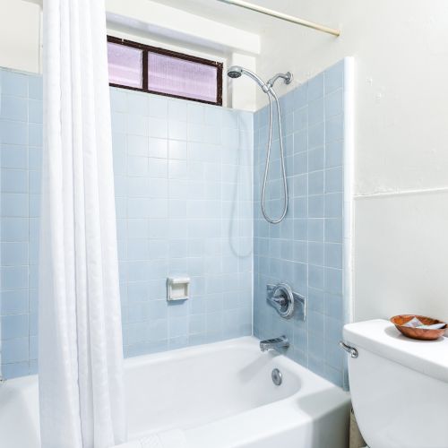 A bathroom with a blue-tiled bathtub and shower, a white curtain, a toilet, and a small window above the shower area is shown.