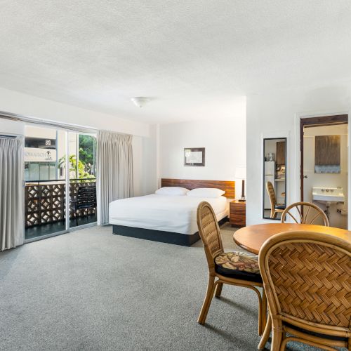 The image shows a hotel room with a bed, a small dining table and chairs, and a TV stand, adjacent to a balcony with curtains.
