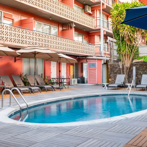 The image shows a hotel pool area with lounge chairs, umbrellas, and a circular pool, surrounded by a building with balconies and tropical plants.