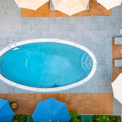 An aerial view of an oval swimming pool surrounded by lounge chairs, umbrellas, and a tiled deck area with greenery.