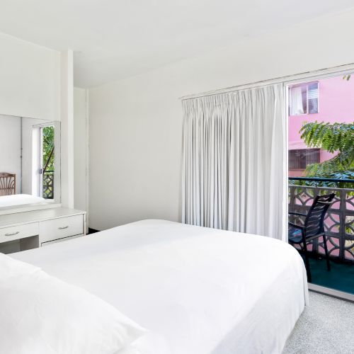 A bright bedroom with a large bed, a vanity, and a balcony view of greenery and a pink building, all in a minimalist style.