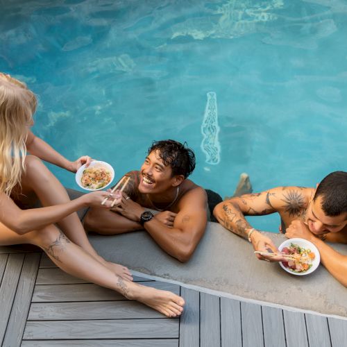 Three people are relaxing by a pool, two in the water and one on the deck, all enjoying food from bowls.