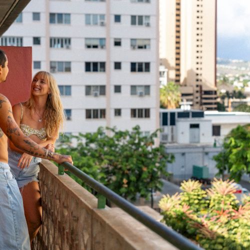 A couple is standing on a balcony, enjoying a sunny day with a view of buildings and greenery in the background.