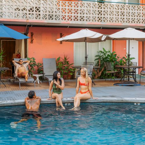 A poolside scene with three people relaxing by the water and another lounging under umbrellas, set against a building with a retro vibe.