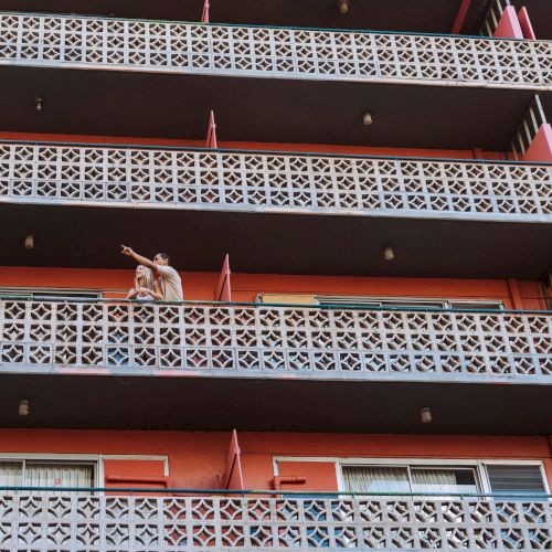 A person stands on a balcony of a multi-level building with geometric patterns and a vibrant orange-red facade.