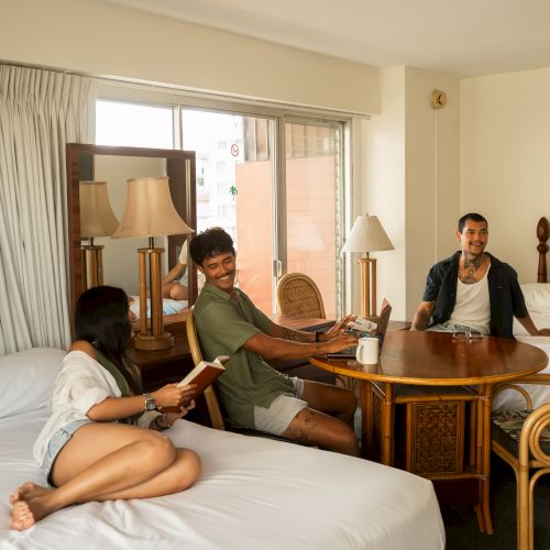 Four people are socializing in a hotel room with two beds, a table, and chairs. The atmosphere is relaxed and casual.