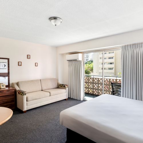 The image shows a hotel room with a bed, sofa, TV, and a balcony. The decor is simple with neutral tones and a view outside.