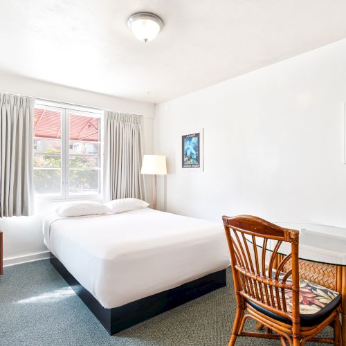 The image shows a bright hotel room with a bed, table, chair, microwave, artwork, and window with curtains.