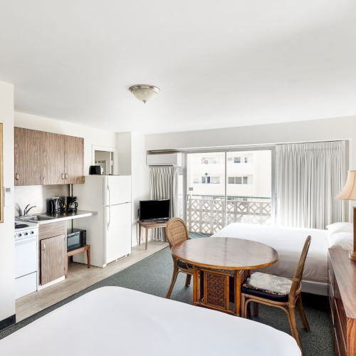 The image shows a bright room with beds, a kitchenette, a table, chairs, a TV, and a lamp, featuring a large window with a balcony view.