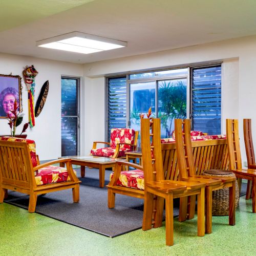 A room with wooden furniture, colorful cushions, decorative masks, and framed art on the wall, next to large windows.