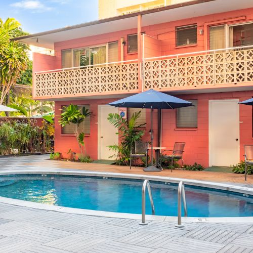 The image shows a courtyard with a small pool, surrounded by a pink building, outdoor furniture, and umbrellas for shade.