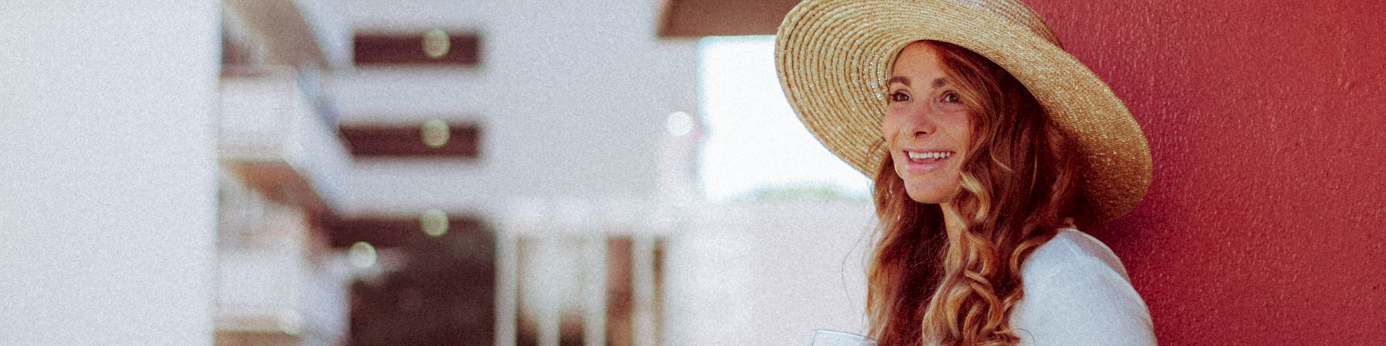 A person in a hat stands on a balcony, smiling, with buildings in the background, creating a relaxed urban scene.