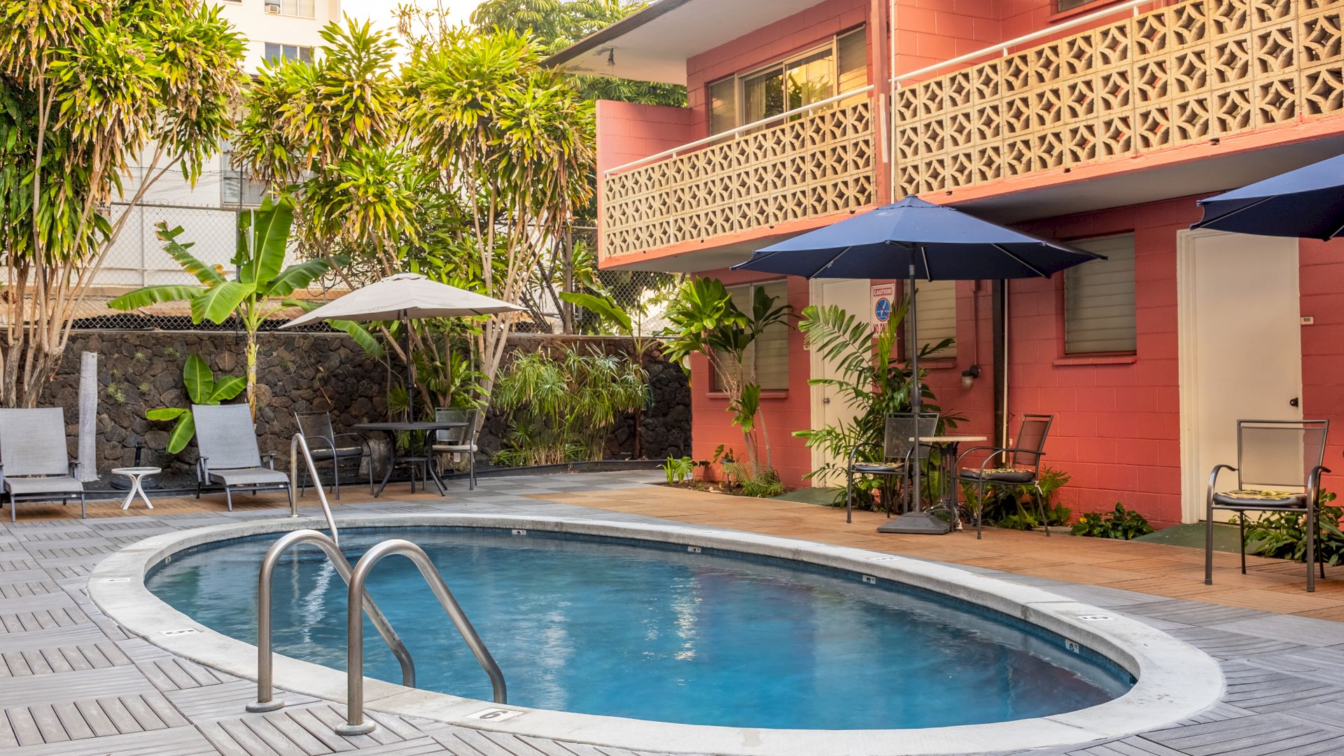 A small, oval pool surrounded by lounge chairs, umbrellas, and tropical plants, next to a building with a patterned railing and pink walls.
